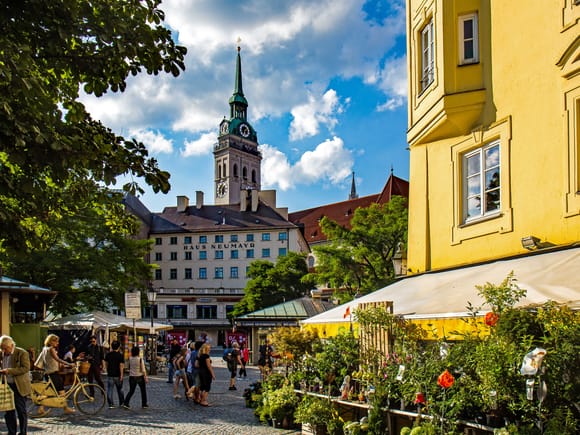 Der Viktualienmarkt München mit Blick auf eine gelbe Gebäudefassade und den Turm des Alten Peters im Hintergrund.