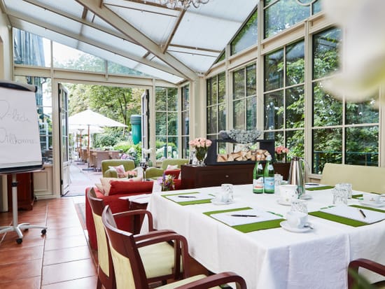 Meeting setup in the light-filled conservatory with a white-clothed conference table, notepads and coffee cups, a flipchart and a view of the garden terrace.