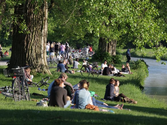 Menschen entspannen und picknicken auf der Wiese im Englischen Garten München, im Hintergrund ein kleiner Bach.