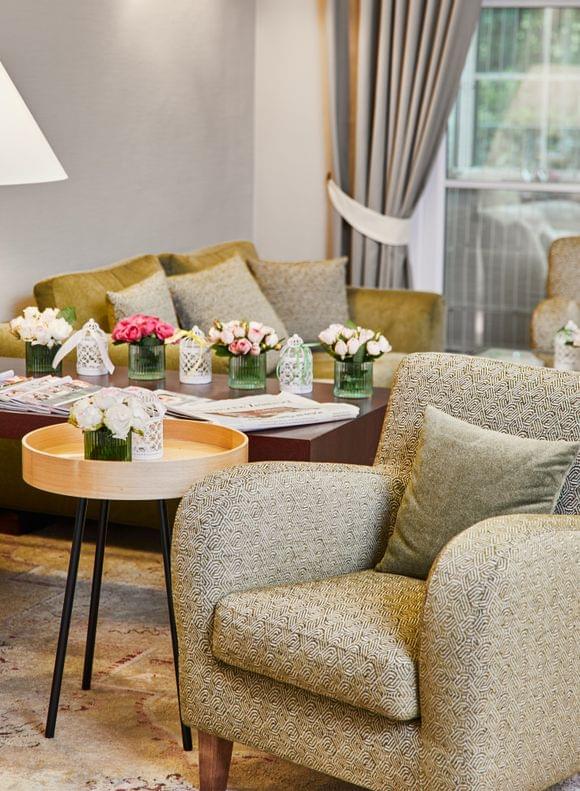 Im Vordergrund steht ein kleiner Holztisch zwischen zwei Sesseln in beige und pastellgrün bezogen, dahinter ein Sideboard mit Zeitschriften und einer angeschalteten Lampe vor einer pastellgrünen Couch mit Dekokissen.
