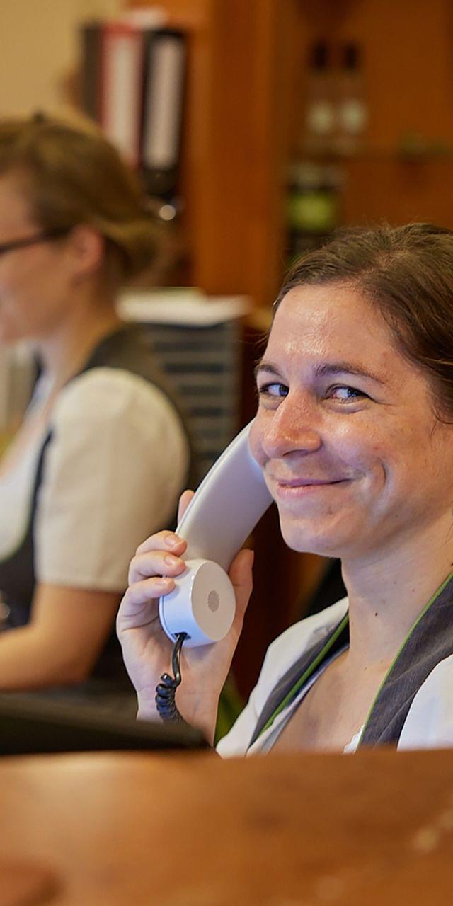 Freundliche Rezeptionistin in Tracht nimmt lächelnd einen Anruf entgegen, im Hintergrund arbeitet eine Kollegin am Computer.