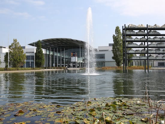 Teich mit Springbrunnen und Seerosen auf dem Gelände der Messe München, im Hintergrund die modernen Gebäude des Messezentrums.
