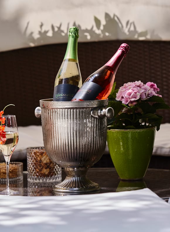Zwei Flaschen Frizzante im Sektkühler mit garnierten Gläsern und Hortensie auf rundem Loungetisch, umgeben von weißen Polstern der gemütlichen Gartenlounge.