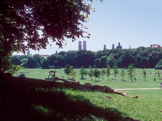 Weite Wiesenfläche im Englischen Garten München mit Blick auf die Türme der Frauenkirche am Horizont, gerahmt von Baumzweigen im Vordergrund.