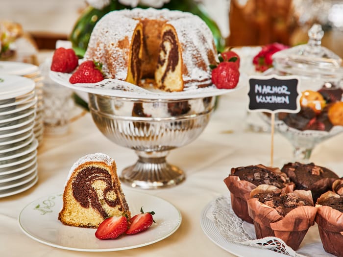 Marble cake dusted with powdered sugar and garnished with strawberries on a silver cake stand, with a served slice in front and chocolate muffins.