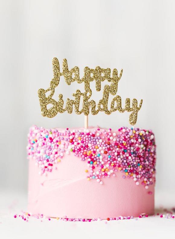 Eine rosa Geburtstagstorte mit bunten Streuseln und goldenem „Happy Birthday"-Topper.