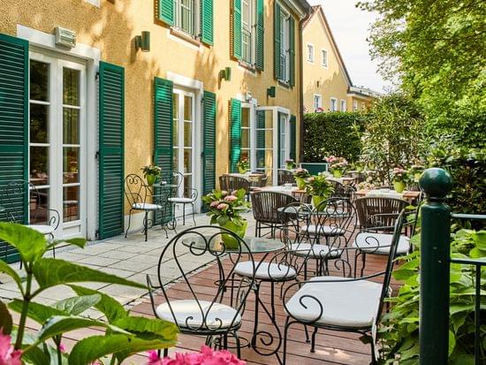 Weitläufige Hotelterrasse mit mehreren Sitzgruppen aus Schmiedeeisen und Holz, üppig blühenden Hortensien und der charakteristischen gelben Fassade mit grünen Fensterläden.