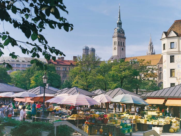 Der Viktualienmarkt München mit bunten Sonnenschirmen über den Ständen und einem Kirchturm im Hintergrund.