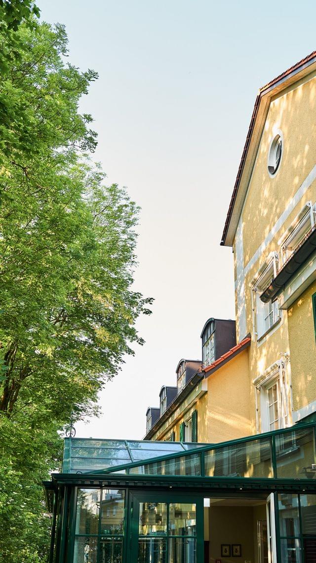 Außenansicht des Hotel Freisinger Hof mit charakteristischer gelber Fassade, grünen Fensterläden und Wintergarten, eingebettet in üppiges Grün.