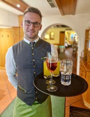 Servicemitarbeiter in bayerischer Tracht serviert ein Tablett mit Aperitif, Orangensaft und Wasser im Restaurant.