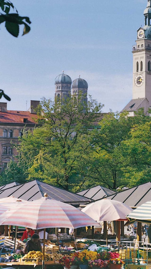 Der Viktualienmarkt München mit bunten Sonnenschirmen über den Ständen und einem Kirchturm im Hintergrund.