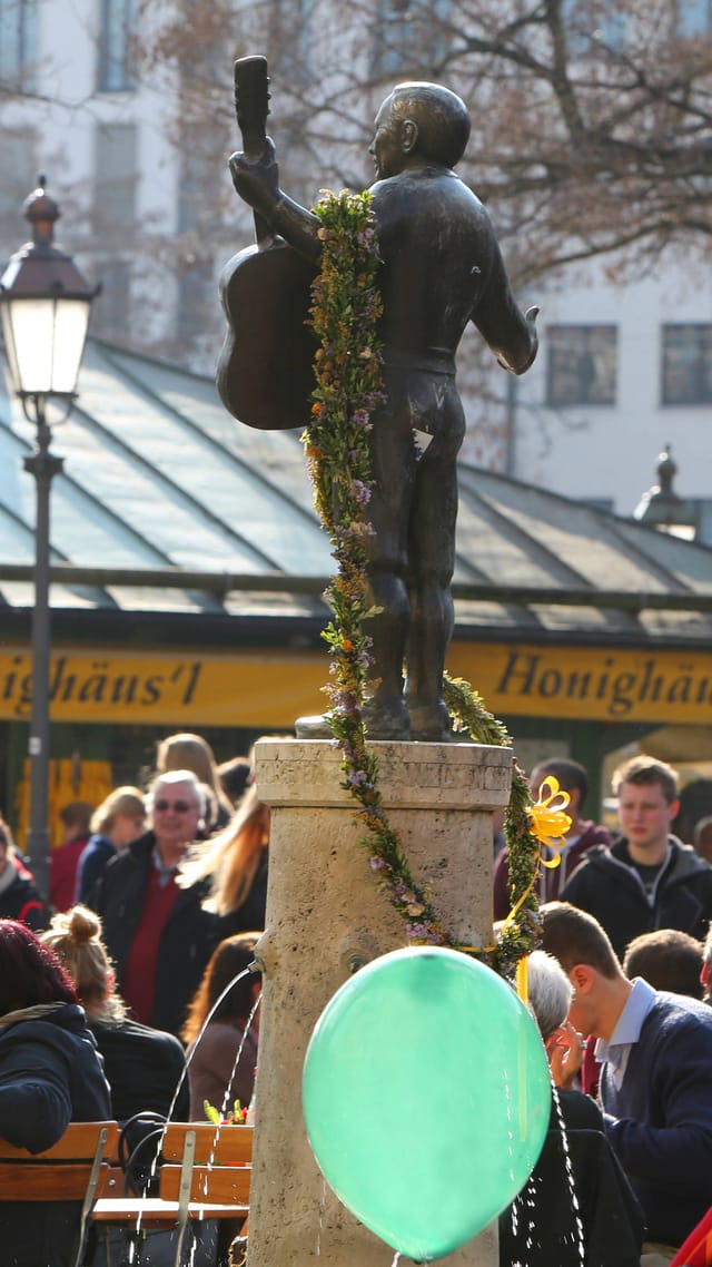Belebter Biergartenbereich auf dem Viktualienmarkt München, Gäste sitzen an Tischen im Freien, im Hintergrund ein Denkmal.