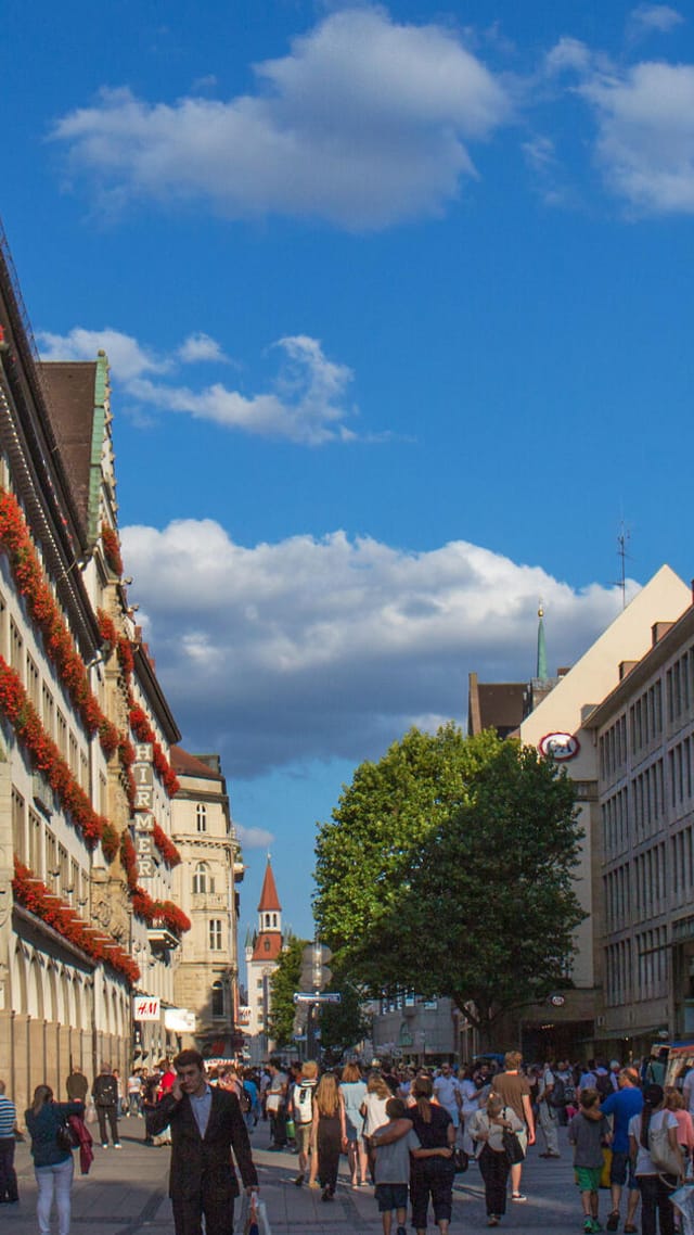 Belebte Fußgängerzone in der Münchner Altstadt mit historischer Fassade und blühenden Balkonen unter blauem Himmel.