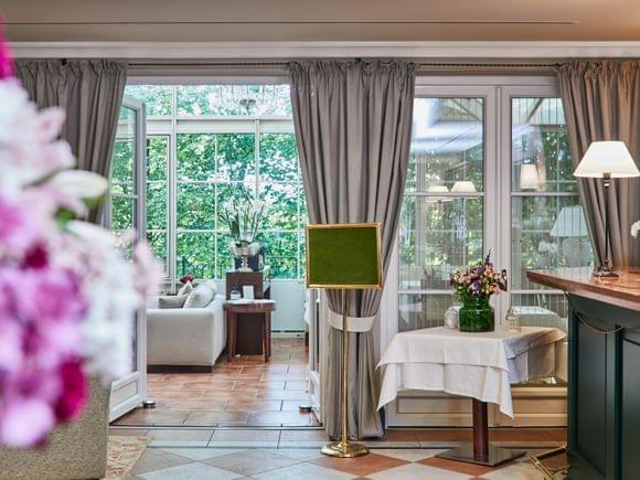 Einladender Durchblick durch elegante Flügeltüren in einen lichtdurchfluteten Wintergarten – im Vordergrund ein gedeckter Tisch mit Blumen, im Hintergrund grüne Bäume und ein gemütlicher Sitzbereich.